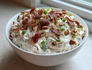 A white bowl filled with a thick, savory cream cheese dip topped with crumbled bacon, chopped pecans, and green onions.