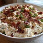 A close-up of a thick, savory cream cheese dip topped generously with crispy bacon crumbles and chopped green onions.