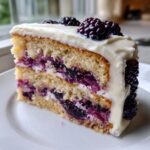 A tall slice of blackberry lavender cake featuring two layers of cake filled with blackberry compote and white frosting, topped with fresh blackberries.