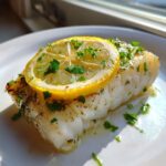 Close-up of a flaky, white chilean sea bass recipe oven baked fillet topped with a lemon slice and fresh parsley.