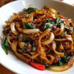Close-up of glossy, wide rice noodles tossed with onions, basil, and chili in a bowl of thai drunken noodles.