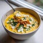 A spoonful of creamy corn soup topped with cotija cheese, cilantro, and crispy tortilla strips.