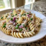 A close-up of creamy Tuna pasta salad featuring rotini pasta, bright green peas, and diced red onion in a white bowl.