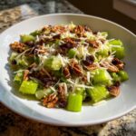 A bright white bowl filled with crunchy celery salad topped with shredded cheese, pecans, and dried cranberries or raisins.