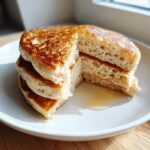 A stack of three fluffy sourdough discard pancakes, cut open to show the airy interior, drizzled with syrup on a white plate.