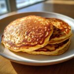 A stack of three perfectly golden gluten free sourdough discard pancakes on a white plate, lit by sunlight.