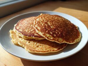 Three golden brown gluten free sourdough discard pancakes stacked on a white plate, lit by sunlight.