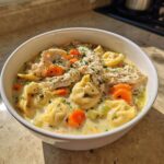 A close-up of a white bowl filled with instant pot chicken tortellini soup, topped with shredded chicken and parsley.