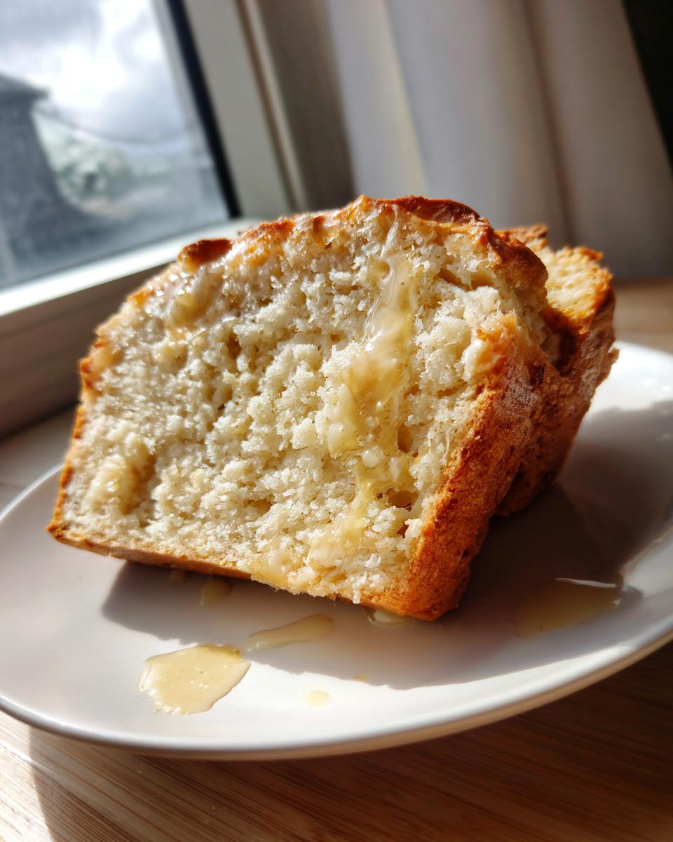 Close-up of a thick slice of moist beer bread drizzled with melted butter or honey on a white plate.
