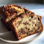 A slice of moist chocolate banana bread packed with chocolate chips, sitting next to the rest of the loaf.