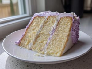 A moist slice of three-layer lavender cake with white and purple frosting on a white plate.