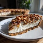 A thick slice of pecan cream pie showing a graham cracker crust, creamy white filling, and a topping of glazed pecans.