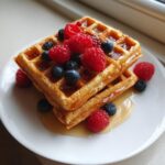 Two stacked protein cottage cheese waffles topped with fresh raspberries, blueberries, and drizzled with syrup.