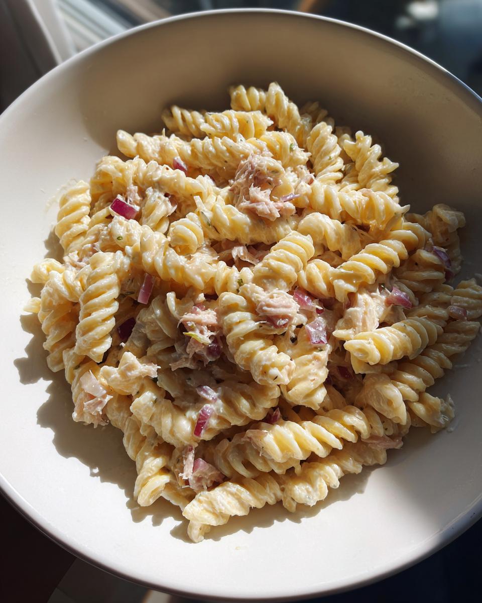 Close-up of creamy protein tuna pasta salad made with fusilli pasta and red onion chunks in a white bowl.