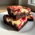 Two stacked, rich chocolate raspberry cheesecake brownies showing swirls of cream cheese and bright red fruit on a white plate.