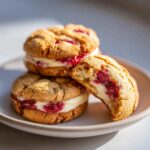 Close-up of stacked raspberry cheesecake cookies, showing creamy filling and bright raspberry jam inside one cookie that has a bite taken.