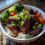 A close-up of a delicious sticky chicken rice bowl topped with glossy sauce, green onions, and sesame seeds.