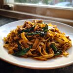 Close-up of steaming street style Thai drunken noodles with wide rice noodles, ground meat, and basil leaves on a white plate.
