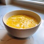 A close-up of a white bowl filled with bright yellow sweet corn soup featuring delicate white egg ribbons.