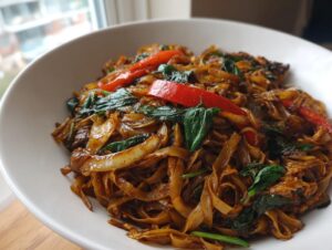 Close-up of a white bowl filled with saucy, dark brown thai drunken noodles, basil, and red peppers.