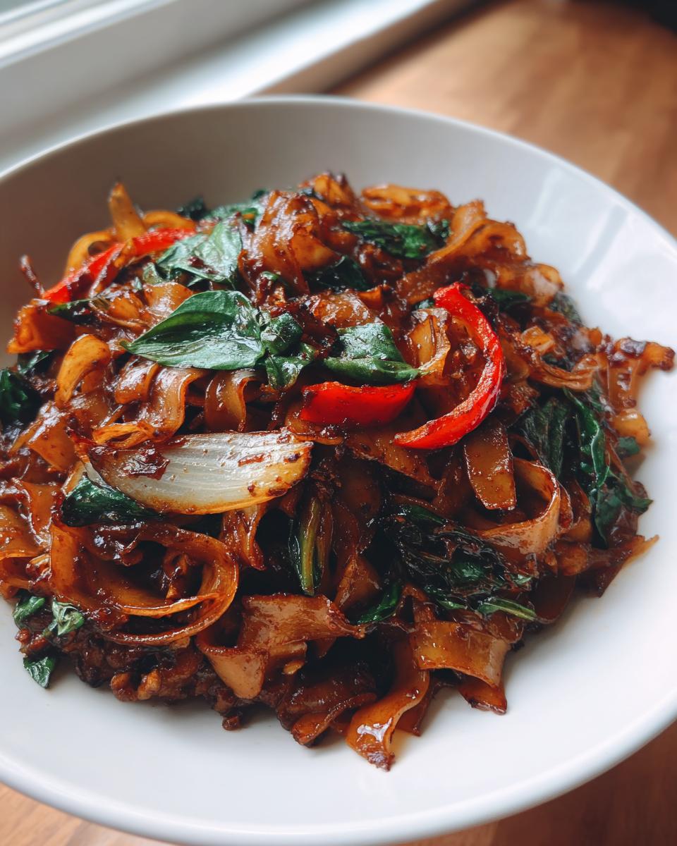 Close-up of glossy, dark brown thai drunken noodles mixed with onions, red peppers, and basil leaves in a white bowl.