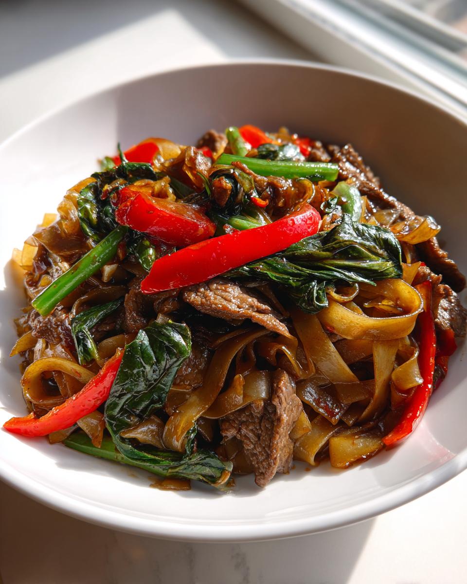 Close-up of a white bowl filled with glossy thai drunken noodles with beef, red peppers, and green vegetables.