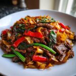 Close-up of a white plate piled high with vibrant thai drunken noodles with beef, peppers, and basil.