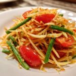 A close-up shot of a vibrant green papaya salad featuring shredded papaya, bright red cherry tomatoes, and green beans.
