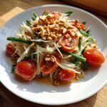 A close-up of fresh papaya salad featuring shredded green papaya, cherry tomatoes, green beans, and crushed peanuts.