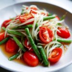 Close-up of a vibrant raw papaya salad featuring shredded papaya, bright red cherry tomatoes, and crisp green beans in a savory dressing.