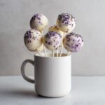 Several white chocolate lavender cake pops decorated with purple sprinkles standing upright in a white ceramic mug.