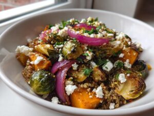 A white bowl filled with a colorful roasted vegetable salad featuring Brussels sprouts, butternut squash, red onion, and feta over quinoa.
