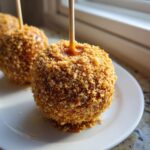 A close-up of one delicious apple pie caramel apples coated in a thick layer of golden crumbs, resting on a white plate.