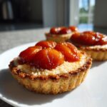 Close-up of a freshly baked apricot-almond tartlet topped with glazed apricot halves and almond frangipane on a white plate.