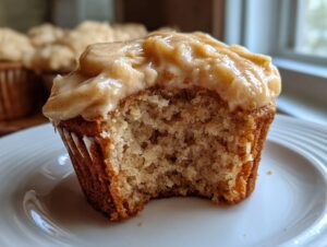 A close-up of a banana cupcake with cinnamon frosting, showing a bite taken out to reveal the moist crumb.