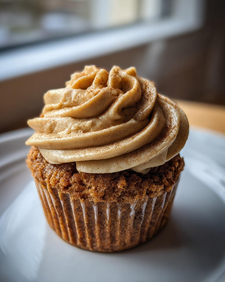 A close-up of a single banana cupcake topped with swirled cinnamon frosting and a dusting of cinnamon spice.