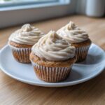 Three moist banana cupcakes from box cake hack, topped with creamy spiced frosting, sitting on a white plate.