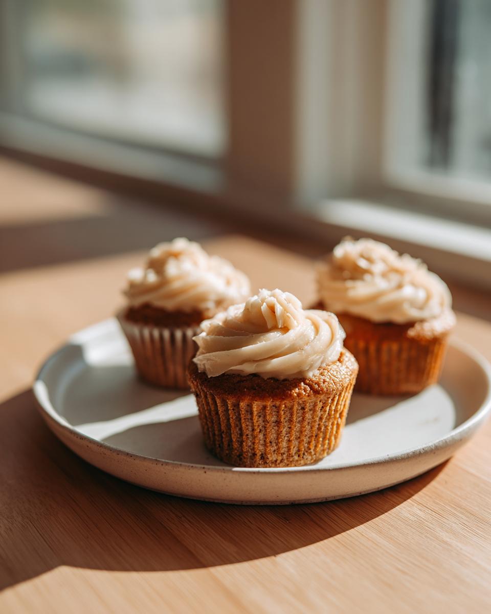 Three frosted banana cupcakes from box cake hack sitting on a light ceramic plate in warm sunlight.