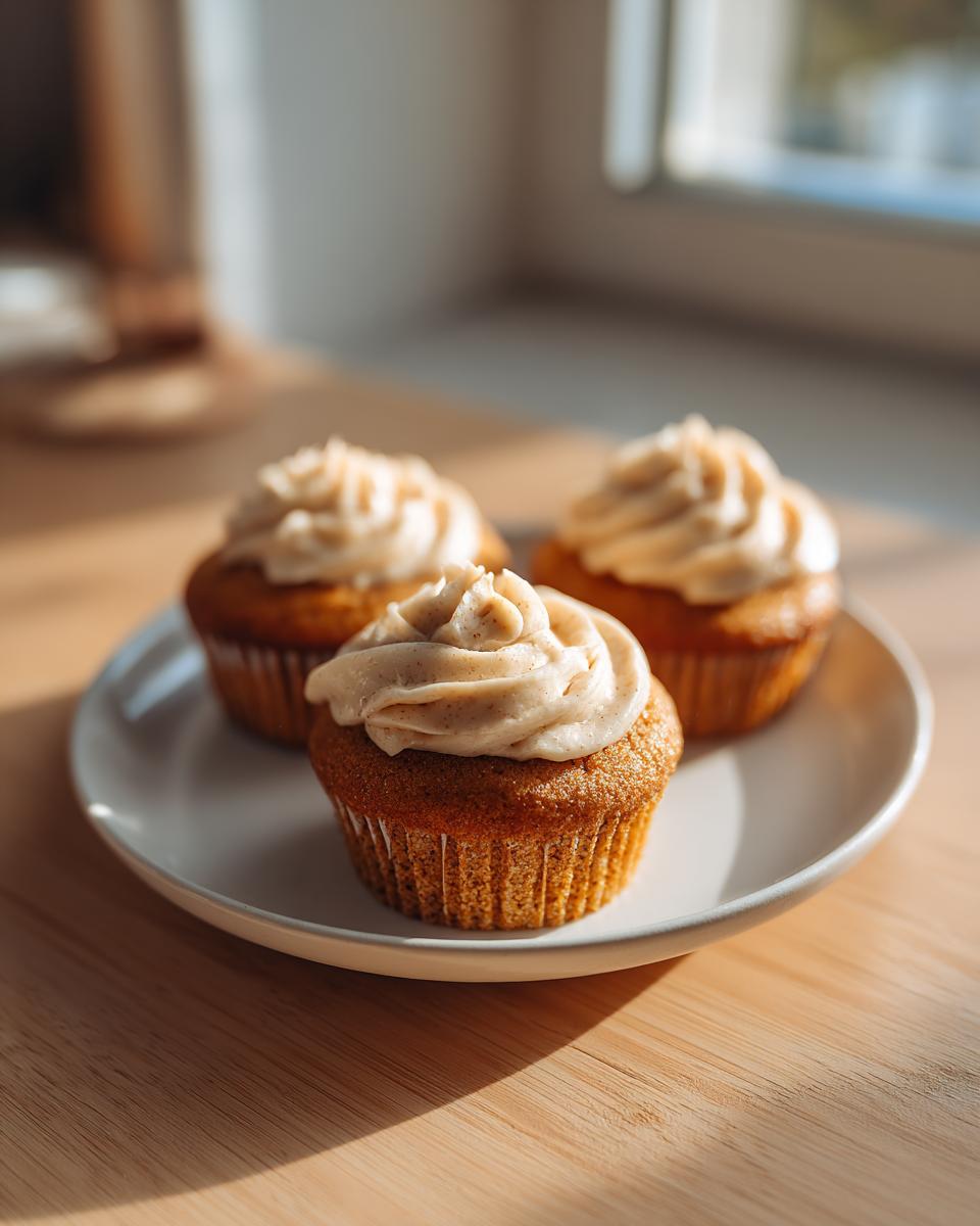 Three moist banana cupcakes from box cake hack topped with cinnamon-flecked frosting on a white plate.