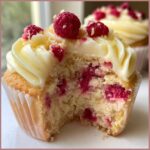 Close-up of a lemon raspberry cupcake with a bite taken out, showing moist cake and fresh raspberries inside, topped with white frosting.