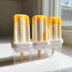 Three freshly made boozy piña colada ice pops with yellow swirls in a white and clear plastic mold on a bright countertop.