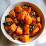 Close-up of perfectly cooked, glazed braised butternut squash cubes topped with crispy sage leaves in a white bowl.