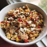 Close-up of a white bowl filled with shredded chicken, corn kernels, diced red peppers, cotija cheese, and sprinkled with paprika, ready to eat chicken street corn salad.