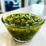 Close-up of chunky, bright green fermented green salsa with visible seeds and herbs in a clear glass bowl.
