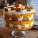 Close-up of a creamy pumpkin trifle dessert layered with orange pumpkin filling, white cream, and cake cubes in a glass pedestal bowl.
