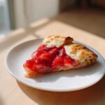 A close-up photo of a single slice of easy strawberry galette with bright red filling on a white plate.