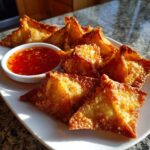 A plate of crispy, golden brown crab rangoons served with a small bowl of bright red sweet chili dipping sauce.