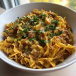 A close-up of a white bowl filled with creamy ground beef goulash mixed with wide egg noodles and topped with fresh parsley.