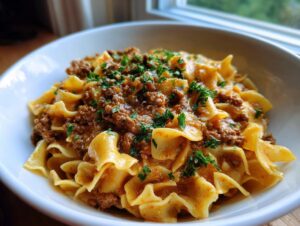 A close-up of a white bowl filled with creamy ground beef goulash served over wide egg noodles and topped with fresh parsley.