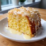 A close-up of a slice of moist Hawaiian wedding cake topped with coconut flakes and a sweet vanilla glaze.